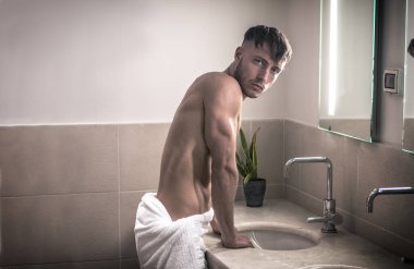 Sabahları banyoda, lavabo ve aynayla kameraya bakan gömleksiz genç bir adam.