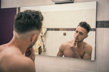 Gömleksiz genç adam sabah banyo aynasında sakalını inceliyor.