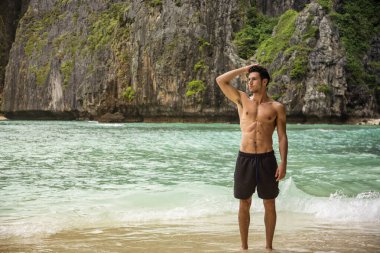 Tayland, Phuket Adası 'nda bir plajda duran yakışıklı genç bir adamın yarı vücut fotoğrafı. Üstsüz, boxer şort giymiş, kaslı fit bir vücut gösteriyor.