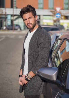 Beyaz gömlekli genç çekici bir adamın portresi şehir caddesinde yeni stil cilalanmış arabasına yaslanmış, kameraya bakıyor.