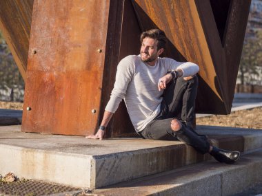 Şehirdeki yakışıklı bir adam Avrupa 'da paslı bir metal yapının yanında oturuyor ve başka tarafa bakıyor.