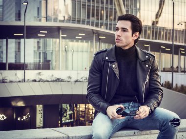 Modern şehirde oturan, siyah deri ceket ve kot pantolon giyen yakışıklı bir genç.