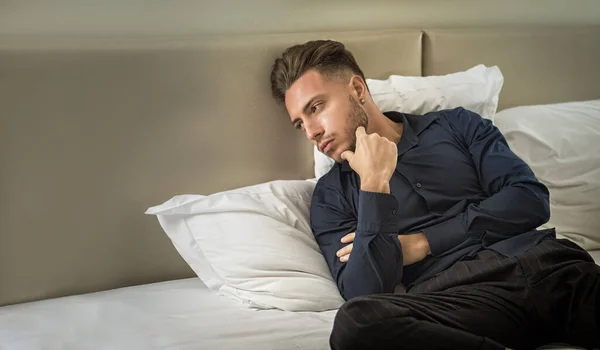 Sexy male model laying alone on bed in his bedroom, looking away to a side with a seductive attitude,, wearing blue elegant shirt