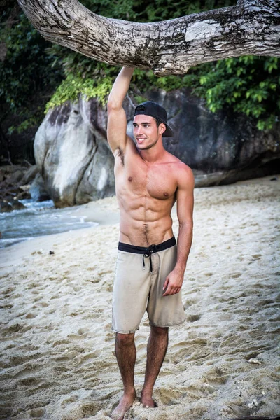 Tayland, Phuket Adası 'nda bir plajda duran yakışıklı genç bir adamın yarı vücut fotoğrafı. Üstsüz, boxer şort giymiş, kaslı fit bir vücut gösteriyor.