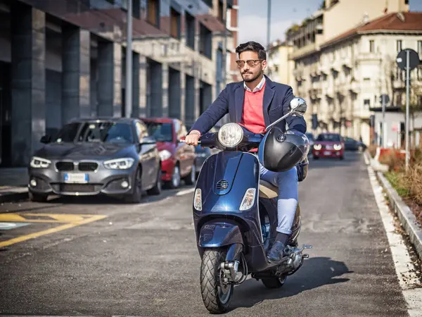 Takım elbiseli ve güneş gözlüklü genç bir adam güneşli şehirde boş bir yolda scooter kullanıyor..