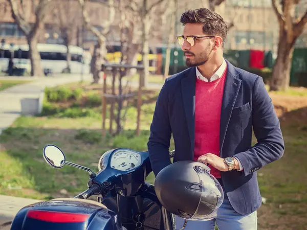 Takım elbiseli ve güneş gözlüklü genç adam güneşli şehirde boş bir yolda klasik İtalyan scooterının yanında duruyor..