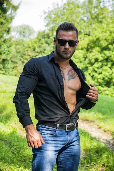 Handsome Hunk Man Outdoor in City Park, Opening his Shirt to Show Muscular Torso and Chest During Daytime, Wearing Black Shirt and Sunglasses