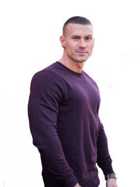 Bir adam düz beyaz bir duvarın önünde duruyor. Yüzünde tarafsız bir ifadeyle doğrudan kameraya bakıyor. Arka plan basit ve minimalistik..