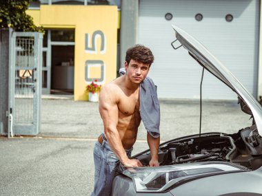 Kaslı yapılı genç bir adam kaputu açık bir arabanın motorunu inceliyor. Kot pantolon giyiyor ve gömleğini omzunun üzerinden tutuyor, şehir ortamında geçiyor..