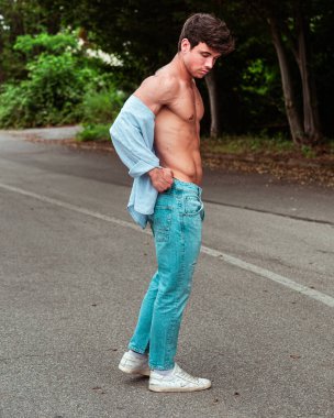 Açık mavi kot pantolon ve kısmen düğmesiz gömlek giyen genç bir adam ıssız bir yolda duruyor. Düşünceli görünüyor, doğada rahat ve rahat bir atmosfer yaratıyor..