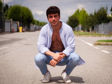 Issız bir sokakta, açık gömlek ve kot pantolon giymiş genç bir adam çömeliyor. Hava açık, yol boyunca yeşil ağaçlar var. Bu rahatlatıcı bir güven anı yakalar..