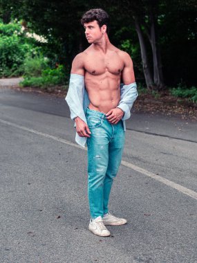 Genç bir adam sakin bir yolda açık mavi kot pantolon ve bol bir gömlek giyerek kendine güvenerek duruyor, kaslı bir fiziği var. Çevredeki yeşillik ana doğal bir zemin katıyor..