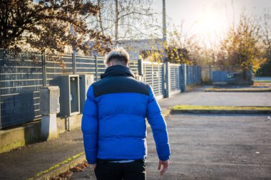 Sarı saçlı ve mavi gözlü genç bir adam mavi ve siyah bir ceketle uzaklaşıyor. Güneş arkasından batıyor, sakin bir şehir bölgesinde sıcak bir atmosfer yaratıyor..