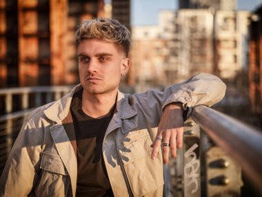 Mavi gözlü genç sarışın bir adam alacakaranlıkta korkuluklara yaslanıyor. Şık kıyafeti ve rahat duruşu şehrin ışıkları parlamaya başladığında kendine güvenini gösteriyor..