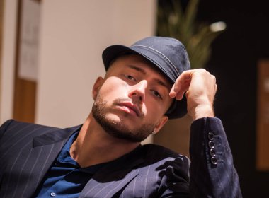 Terzi kıyafeti giymiş ve fötr şapka takmış bir adam arkasına yaslanmış, karizma ve güven saçıyor. Zarif iç mekan dekoru, onun cilalı görünümünü ve rahat tavırlarını geliştirir..