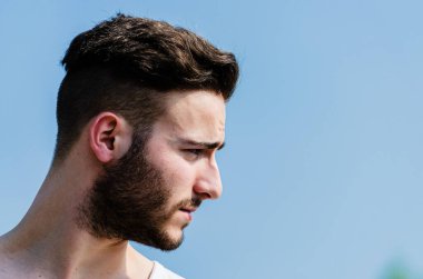 Genç bir adam dışarıda dikiliyor, düşünceli bir şekilde yan tarafa bakıyor. Kuvvetli profili açık mavi gökyüzü tarafından vurgulanmış, güneşli bir günde derin düşüncelere daldığını ya da yansıdığını gösteriyor..