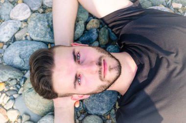 Genç bir adam plajda pürüzsüz, yuvarlak taşlara uzanıp güneşli bir günde güneşin tadını çıkarıyor. Onun gevşek ifadesi doğayla bir huzur ve huzur hissi veriyor..