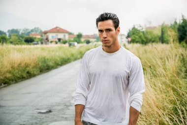 Bir adam sessiz bir kırsal yolda uzun kollu ıslak bir gömlek giyerek tek başına duruyor. Çevresindeki çimenler uzun ve binalar arka planda bulutlu bir gökyüzünün altında görünüyor..