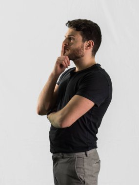 Siyah gömlek ve açık renk pantolon giymiş, dudak dudağa dokunmuş, düşünceli bir ifadeyle genç bir adam profilde duruyor. Arka plan bembeyaz, dalgın duruşunu vurguluyor..