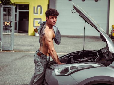 Kaslı yapılı genç bir adam sanayi bölgesinde bir araba motoru üzerinde çalışıyor. Üstsüz ve aracı tamir etmeye odaklanmış durumda. Arka planda sarı duvarlar ve bir garaj var..