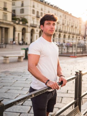 Gün batımında kalabalık bir şehir meydanındaki korkulukların yanında duran genç bir adam. Basit bir tişört giyer ve gün sona ererken düşünceli bir şekilde uzaklara bakar..