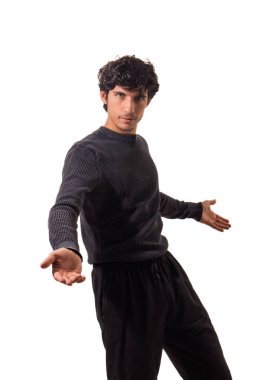 Siyah kazaklı ve siyah pantolonlu bir adam, tarafsız bir ifadeyle dosdoğru karşıya bakıyor. Sade, kapalı bir ortamda asgari ışıklandırmayla duruyor gibi görünüyor..