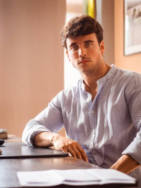 Parlak ışıklandırılmış odada düşünceli bir şekilde masada oturan genç bir adam görülüyor. Çalışırken ya da ders çalışırken, etrafını çevreleyen düzenli bir çalışma alanı ile odaklanmış görünüyor..