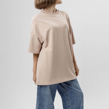 Açık bej renkli kısa kollu tişörtlü ve mavi kotlu kadın iç mekanda minimalist duruyor..