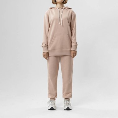 Bej kapüşonlu, eşofmanlı, spor ayakkabılı, kapalı mekândaki minimalist stüdyosunda duran bir kadın..