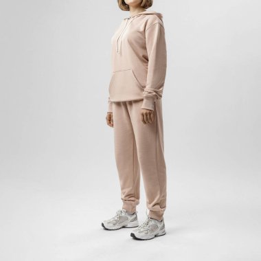 Bej kapüşonlu ve eşofmanlı, spor ayakkabılı parlak minimalist stüdyoda poz veren bir kadın..