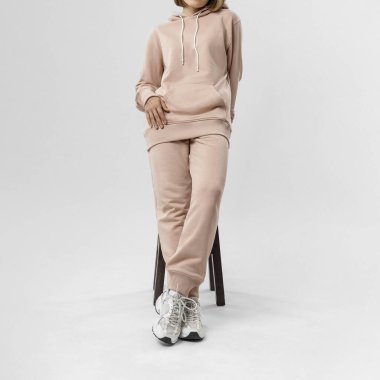 Bej kapüşonlu ve eşofmanlı genç bir kadının parlak bir stüdyoda dikilip günlük kıyafetlerini sergilemesi..