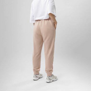 Minimalist bir stüdyo ortamında bej eşofman altı ve beyaz spor ayakkabılar giyen bir kadının arka görünüşü, gündelik rahatlık sergiliyor..