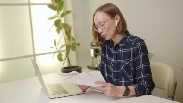 Mulher Freelancer Camisa Azul Óculos Auriculares Examinando Papelada Com Laptop — Vídeo de Stock