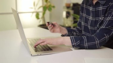 Ekose gömlekli bir kadın ofis ortamında dizüstü bilgisayarına kredi kartı bilgilerini giriyor. Yüksek kalite 4k görüntü.