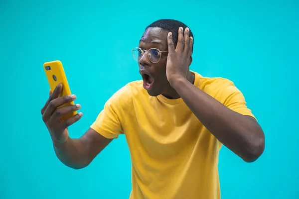 Mutlu heyecanlı Afroamerikalı adam akıllı telefonu açık ağızla tutuyor. Mavi arkaplan üzerinde sarı tişörtle afiş tasarımı için fotokopi alanı var. Vay be, erkek. Arkadaşlarla çevrimiçi oyun