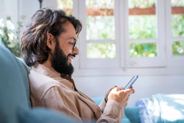 Çekici, gülümseyen, sakallı genç bir adamın portresi oturma odasında telefon kullanarak oturuyor. Mutlu erkek kanepede rahatlar akıllı telefon ekranı mesajlarına bakar ya da internetten mesajlaşır. Teknoloji
