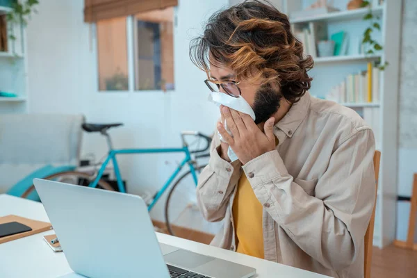 Unhealthy young modern man feeling unwell, sick with running nose. Male manager or freelancer sit at the desk at home office, working distantly, using paper tissue, sneezing, flu, cold.
