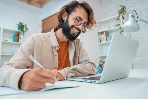 Dizüstü bilgisayarda çalışan başarılı gülümseyen, konferans sırasında notlar alan, internet üzerinden bir webinar dinleyen ve çalışma alanında bilgisayar kullanan bir adam. Yüksek kalite fotoğraf