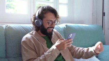 Kulaklıklı neşeli adam kanepede oturup müzik dinliyor, mutlu, rahat bir milenyum adamı huzurlu bir ruh halinde stres olmadan eğleniyor. Yüksek kalite 4k görüntü