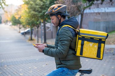 Şehirdeki güneşli caddede bisiklet süren müşterilere yiyecek getiren bir adam. Genç kurye bir sonraki siparişi için akıllı telefonunu kontrol ediyor. Yüksek kalite fotoğraf