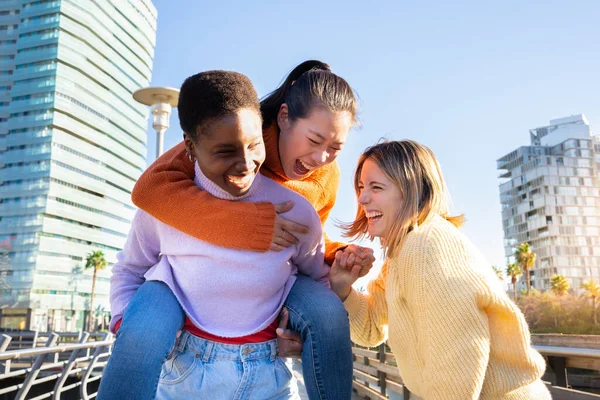 Happy multiracial young women embracing each other walking and piggybacking a friend on city street. Smiling, enjoying and having fun together on sunny day. High quality photo
