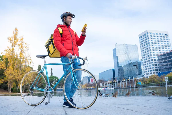 Young smiling delivery rider man at street checking the street address at smartphone app. Outdoor courier with backpack and bike. High quality photo