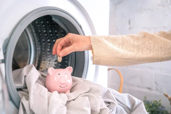 Reduce household bills expenses money savings, hand insert coin in piggy bank. Energy and electricity crisis, washing machine in the background