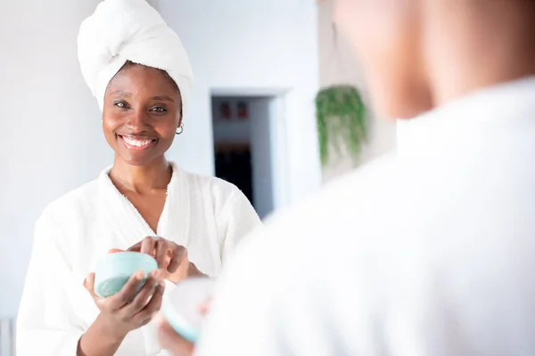 Portrait of happy woman applying natural cosmetic cream, smiling looking in mirror. Skincare routine grooming herself after shower in bathroom
