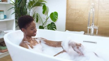 Gülümseyen genç bir kadın köpük banyosundan, cildini sabunla yıkamaktan hoşlanır. Kişisel bakım spa zamanı ve güzellik, vücut farkındalığı. Yavaş çekim kamerası kırmızı yüksek kalite 4K görüntü
