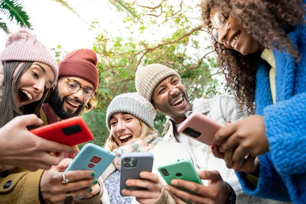 Sosyal medya içeriğini açık havada akıllı telefondan paylaşan bir grup mutlu etnik arkadaş. Kışın kıyafet giyen öğrenciler online oyun oynayan cihazları kullanarak eğleniyorlar..