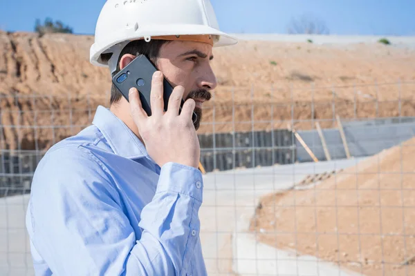 Civil engineer inspecting work calling to supervisor with smartphone standing outdoors at construction site with licenses and blueprints, control, inspect. Copy space for text. High quality photo