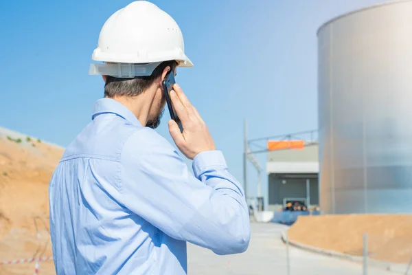 Civil engineer inspecting work calling to supervisor with smartphone standing outdoors at construction site with licenses and blueprints, control, inspect. Copy space for text. High quality photo