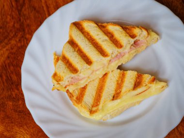 Ahşap masadaki beyaz tabakta ızgara jambon ve peynirli tost.