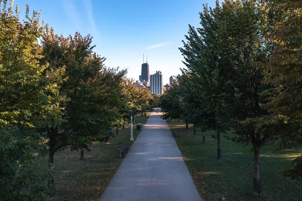 People walk down a paved path lined with trees and park benches in the Lincoln Park neighborhood with a view of downtown high rise buildings and a blue sky above.